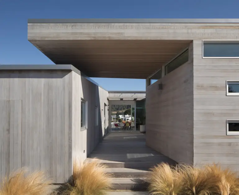 A house with a cantilevered roof that forms a breezeway canopy is architect Cass Calder Smith’s version of a bleached wood beach “shack” for a surfing family.