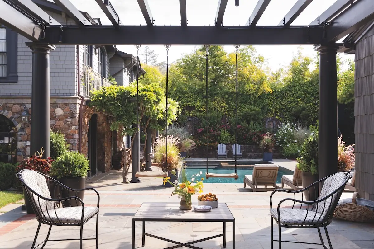 An outdoor patio under an awning with swings hanging from it and a pool and building in the background.