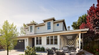 A modern two-story home with cream siding and black accents, surrounded by greenery and featuring a welcoming front yard with outdoor seating and a firepit.