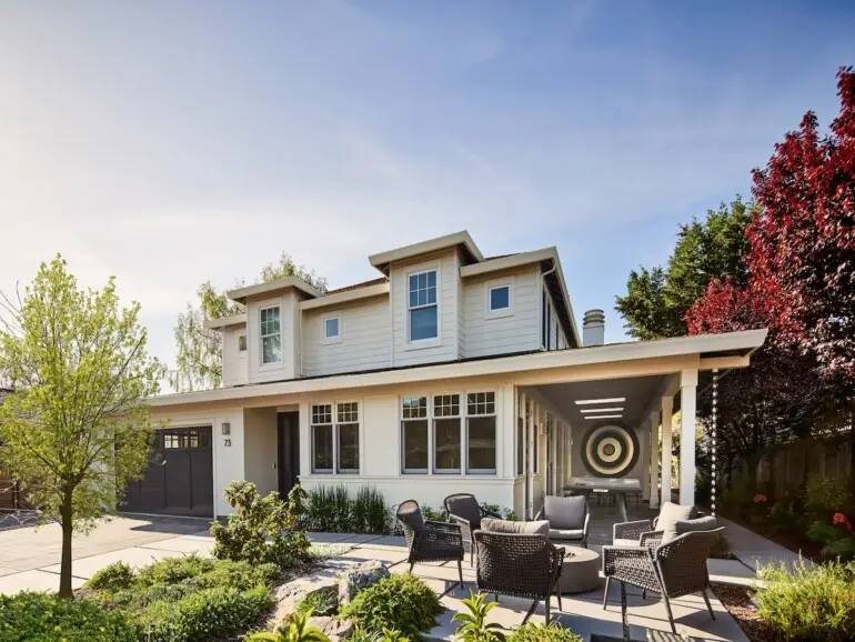 A modern two-story home with cream siding and black accents, surrounded by greenery and featuring a welcoming front yard with outdoor seating and a firepit.