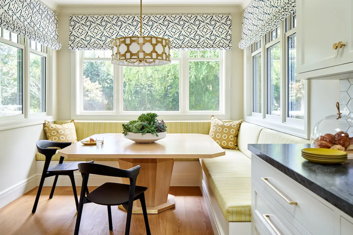 A sunny corner breakfast nook with striped yellow cushions, a light wood table, patterned roman shades, and surrounding windows.