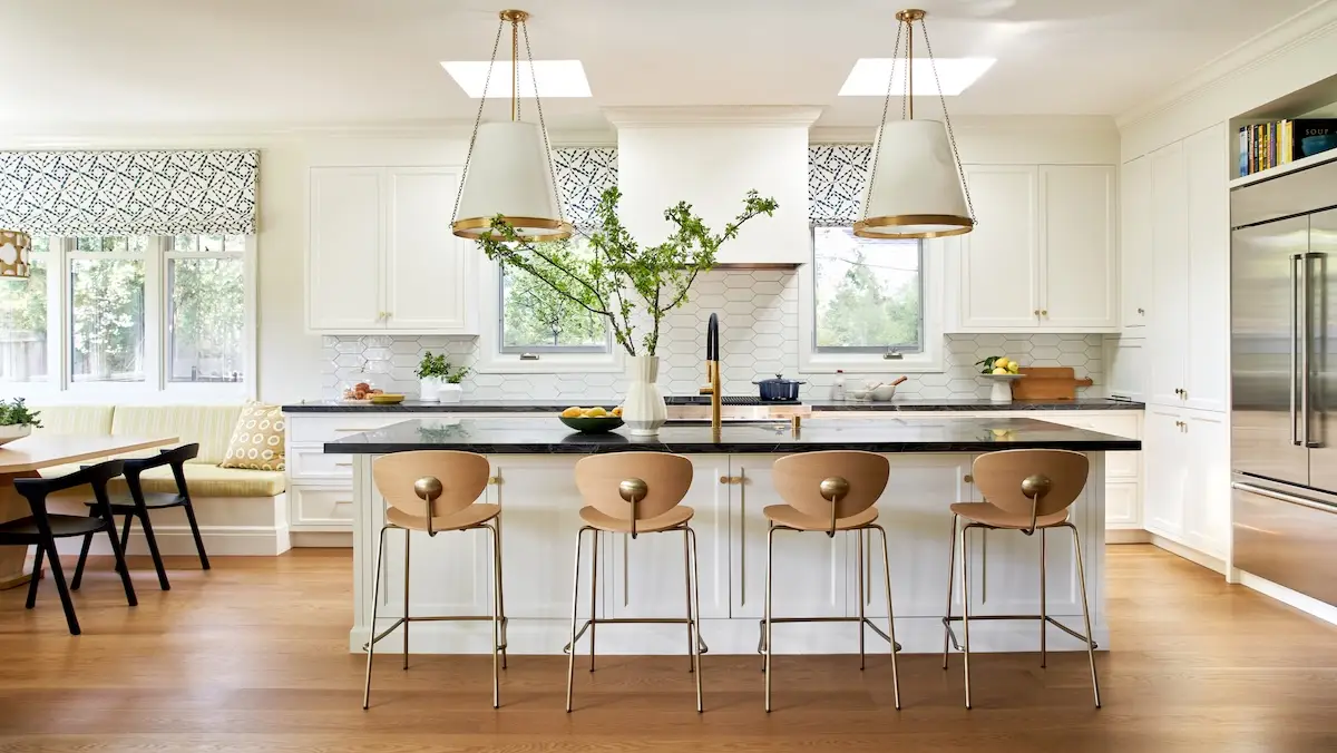 A spacious kitchen featuring a large island with modern wood-and-brass bar stools, white cabinetry, and pendant lights under skylights.