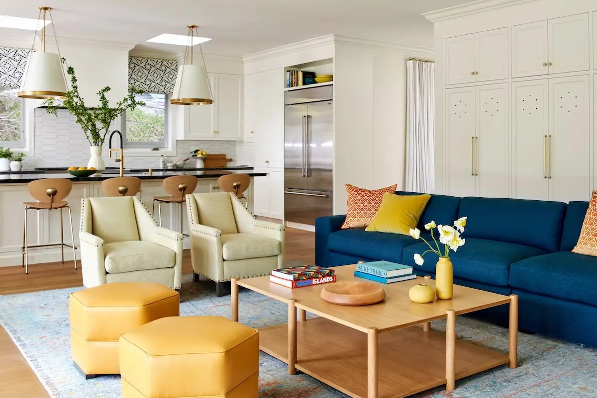 A lively family room with a blue couch, yellow ottomans, and a mix of colorful decor, seamlessly connected to the adjacent kitchen area.