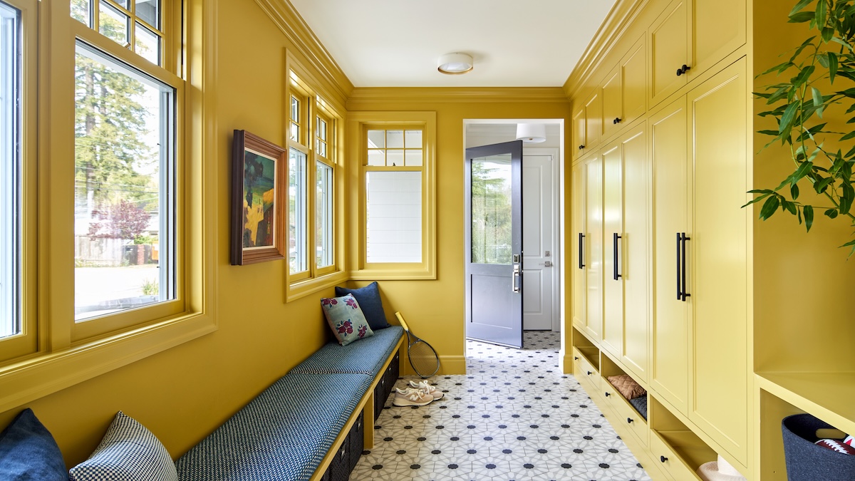 A cheerful, yellow-painted mudroom with built-in cabinetry, a cushioned bench under a row of windows, and patterned tile flooring leading to a front door.