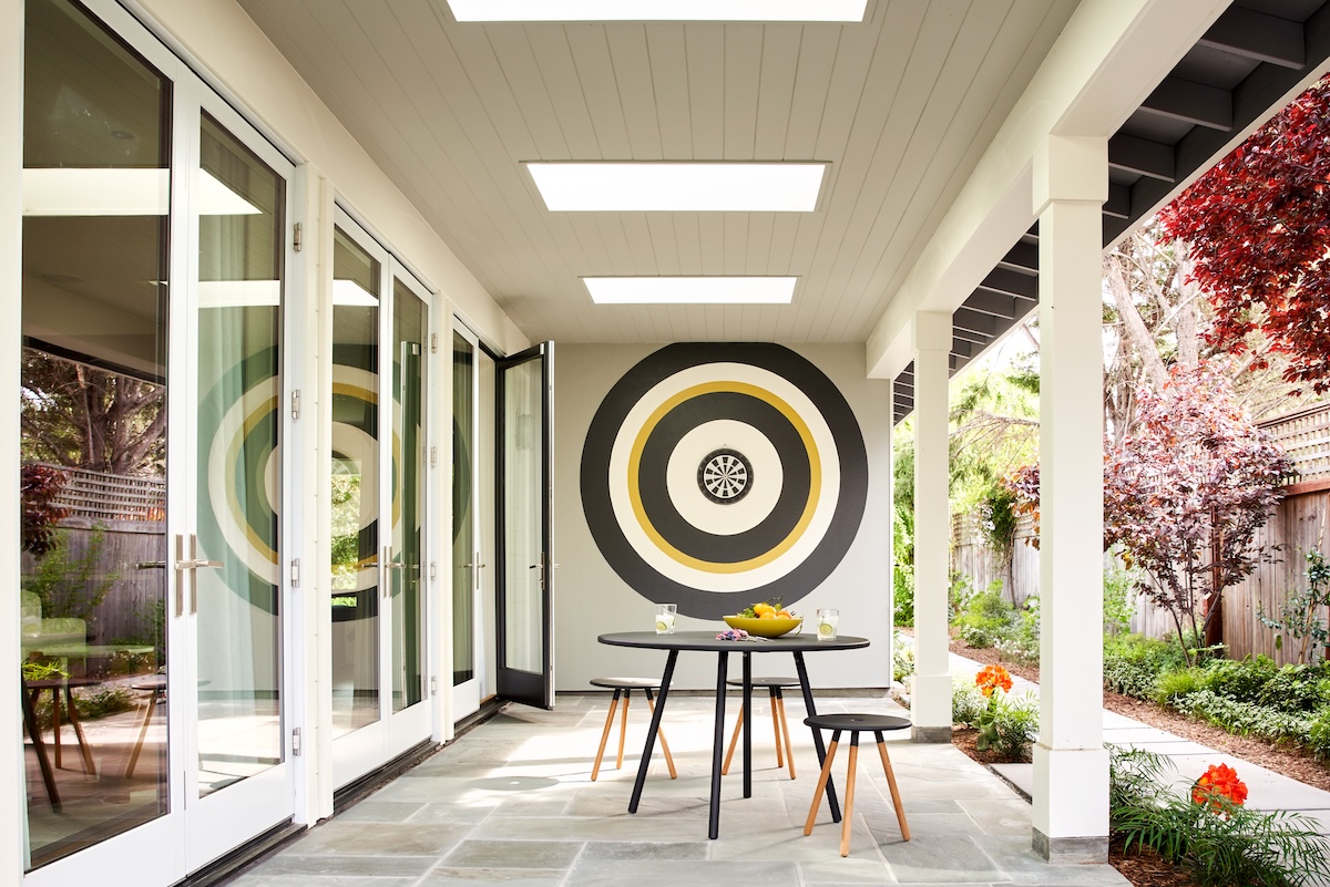 A stylish covered porch with skylights, a round table and stools, and a bold black, white, and yellow concentric-circle mural resembling a dartboard.