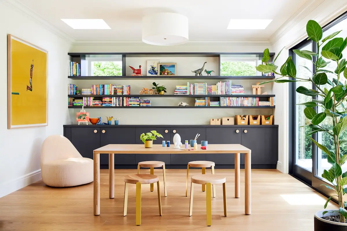 A vibrant playroom featuring a wall of colorful books on dark shelves, a light wood table with stools, and modern art and toys throughout.