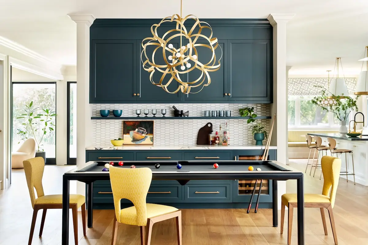 A striking space with a dark teal bar wall, yellow chairs, and a sleek pool table that doubles as a dining table, all under a sculptural gold chandelier.