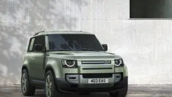 A sage green metallic Land Rover Defender car pictured with a concrete wall in the background.