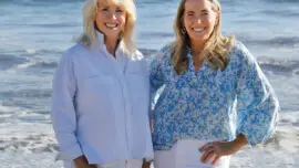 Heather Sandy Hebert and Chase Reynolds Ewald stand side by side smiling at the camera, with waves crashing behind them on a sunny California beach.