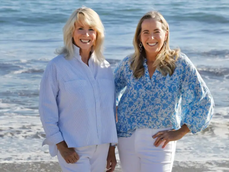 Heather Sandy Hebert and Chase Reynolds Ewald stand side by side smiling at the camera, with waves crashing behind them on a sunny California beach.