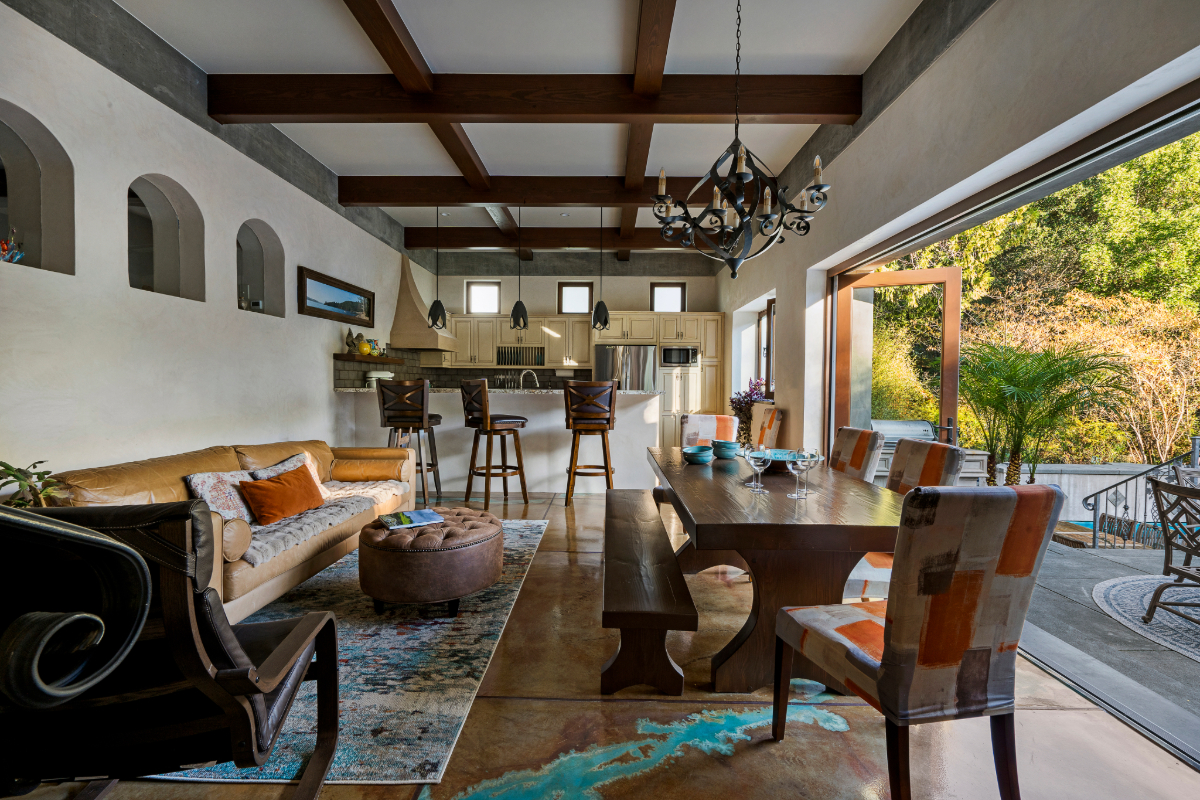 The ADU’s Mediterranean-inspired living and dining area with exposed wood beams, open bi-fold doors to the patio, and a custom wood dining table.