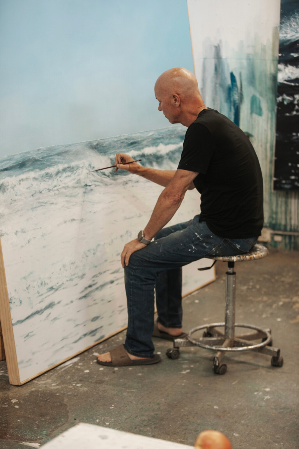 Eric Zener painting a realistic seascape in his studio, sitting on a stool with a brush in hand, focused on fine details of an ocean wave.