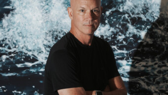 Artist Eric Zener standing with arms crossed in front of a vivid, large-scale ocean wave painting.