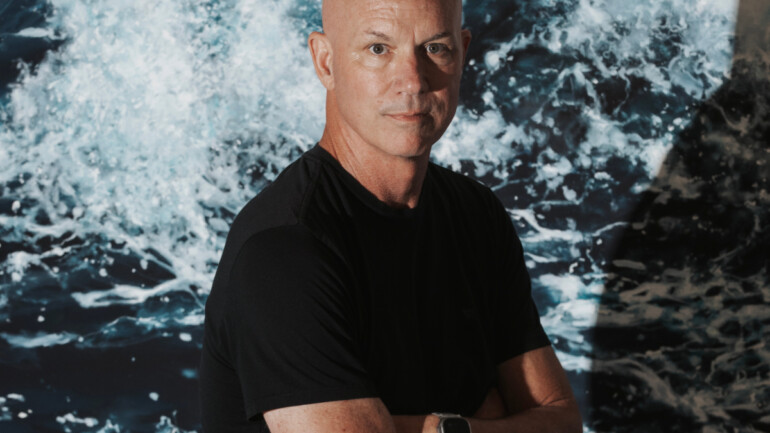 Artist Eric Zener standing with arms crossed in front of a vivid, large-scale ocean wave painting.