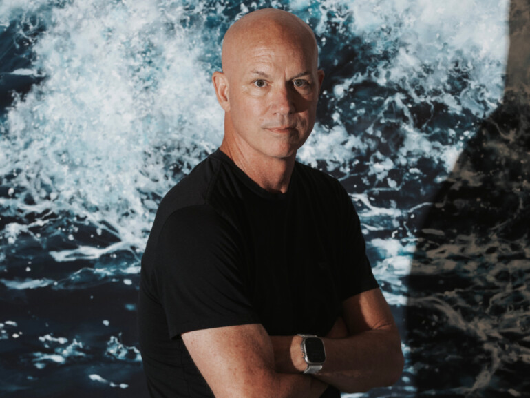 Artist Eric Zener standing with arms crossed in front of a vivid, large-scale ocean wave painting.