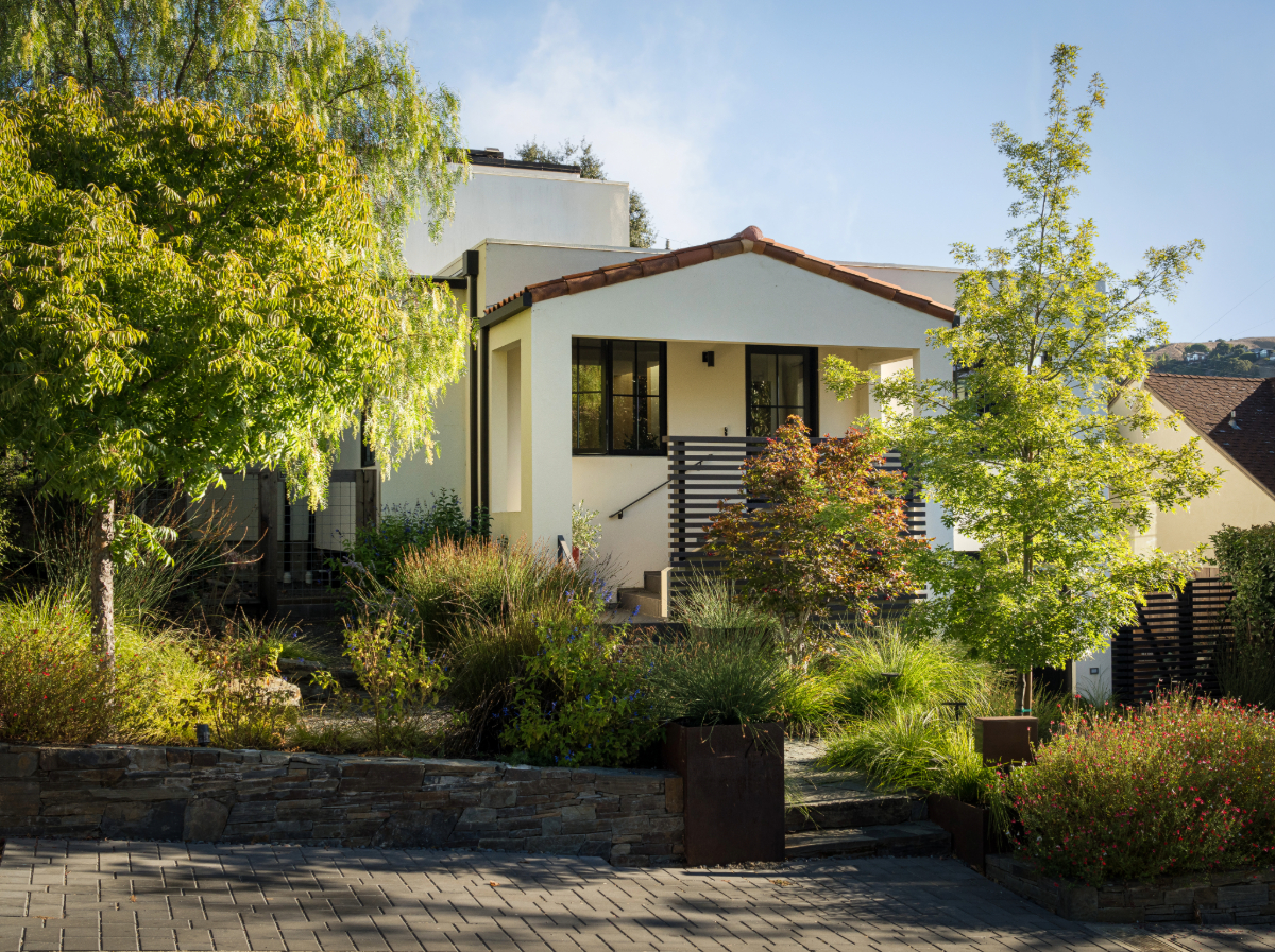 Front exterior of the renovated San Anselmo Spanish-style cottage featuring a red-tiled roof, white stucco façade, black-framed windows, and a lush landscaped front yard with mature trees and greenery.