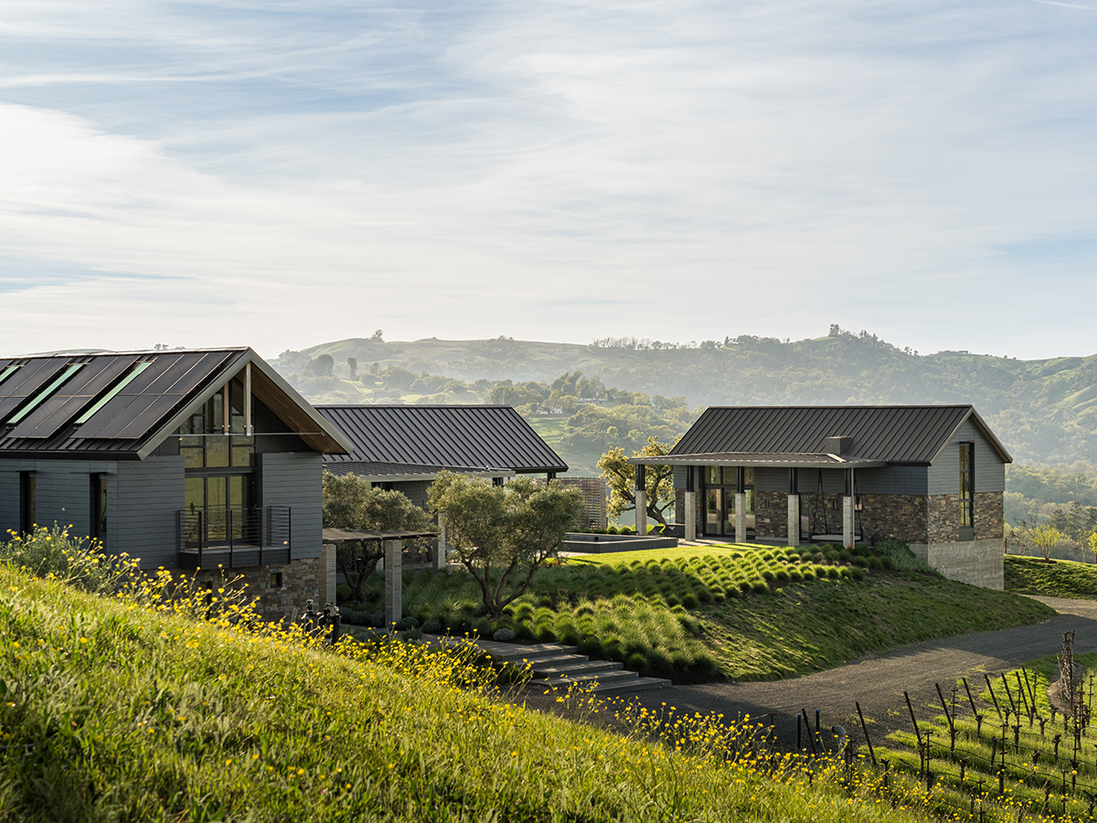 A house perched on a hill, overlooking a lush vineyard in the background under a clear blue sky.