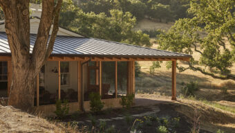 A house with a metal roof perched on a grassy hill, surrounded by trees and a clear blue sky.