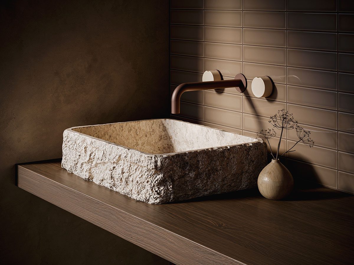 Rectangular carved stone vessel sink on a floating wood vanity with a wall-mounted faucet, set against warm neutral tile and plaster walls