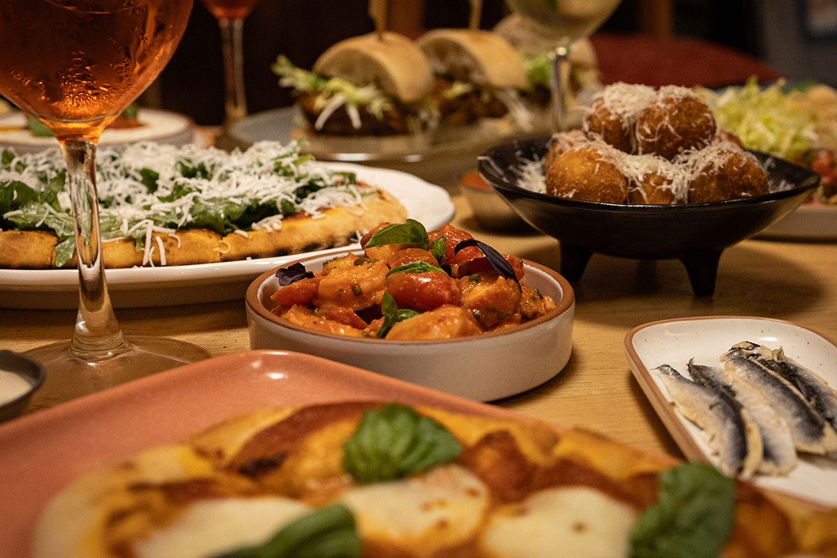 A table full of Italian dishes, including pizza, bruschetta, arancini, sardines, and sliders, accompanied by a glass of Aperol spritz, conveying a cozy, inviting feast.