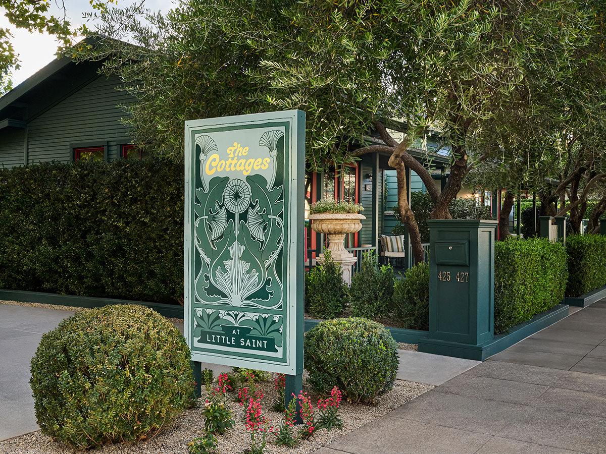 A sign for "The Cottages at Little Saint" is surrounded by lush greenery and a quaint wooden building. The scene feels serene and inviting.