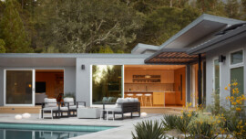 Modern house exterior with large windows, patio seating, and a pool. Warm indoor lighting creates a cozy atmosphere, surrounded by lush trees.