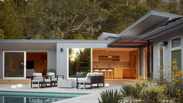 Modern house exterior with large windows, patio seating, and a pool. Warm indoor lighting creates a cozy atmosphere, surrounded by lush trees.