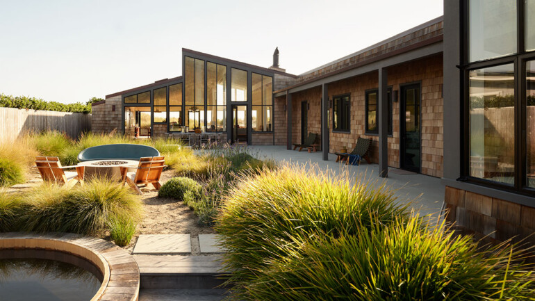 Modern house with large glass windows and wooden shingles, surrounded by lush grasses. A patio with chairs and a hot tub creates a serene, inviting setting.