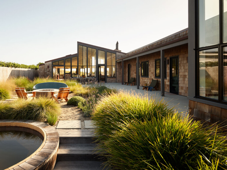Modern house with large glass windows and wooden shingles, surrounded by lush grasses. A patio with chairs and a hot tub creates a serene, inviting setting.
