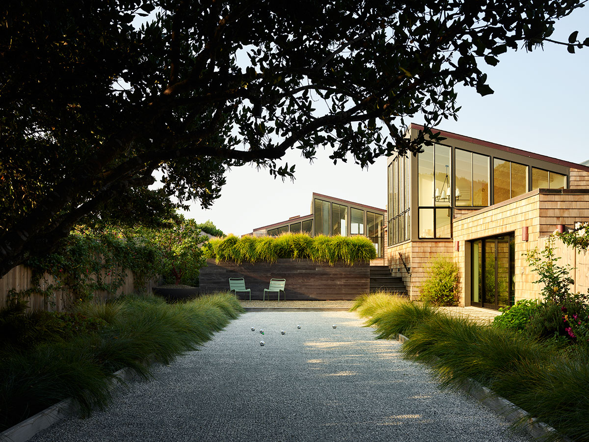 Modern house with large windows surrounded by lush greenery, viewed from a gravel pathway. Two chairs sit in front of a raised garden, evoking tranquility.