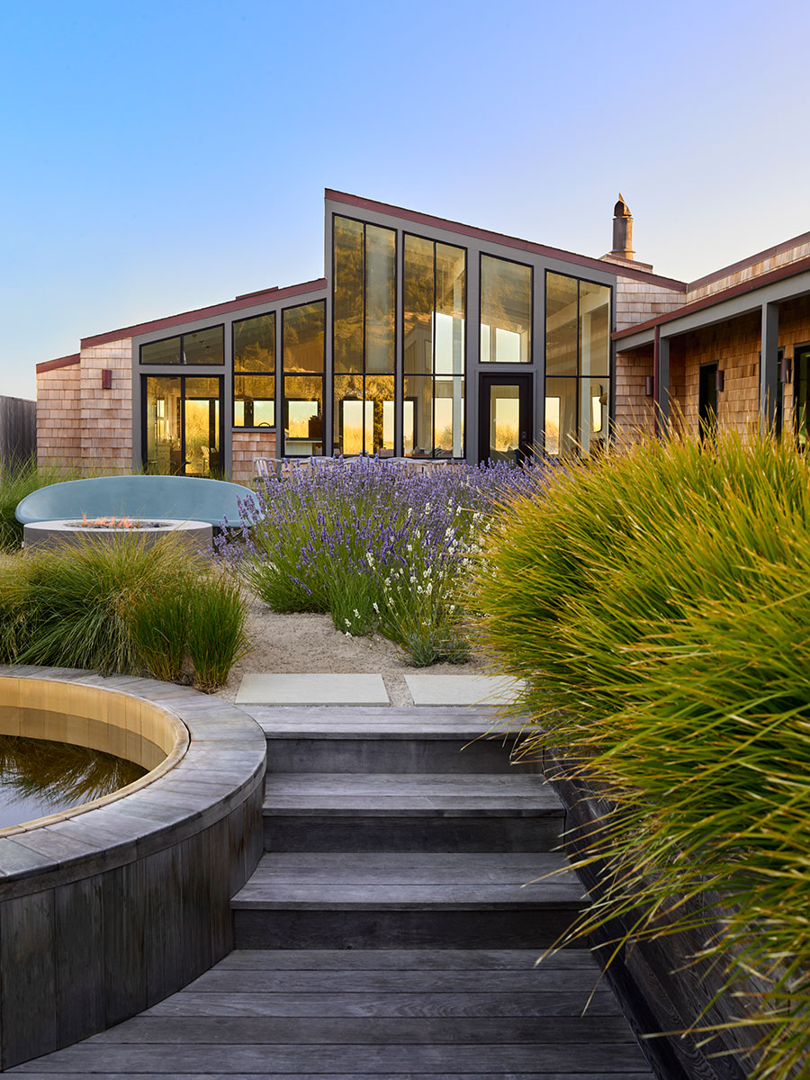 Modern house with large glass windows, surrounded by lush green plants and lavender bushes. Wooden steps lead to a circular pond. Tranquil atmosphere.