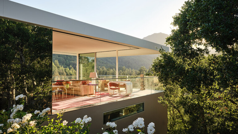 Modern glass-walled living room with pink decor overlooks lush forest, surrounded by white flowers and trees, exuding tranquility and elegance.
