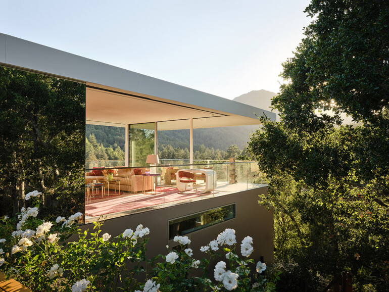 Modern glass-walled living room with pink decor overlooks lush forest, surrounded by white flowers and trees, exuding tranquility and elegance.
