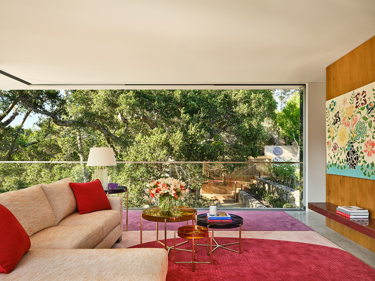 Modern living room with large windows revealing lush greenery. Beige sofa with red cushions, colorful flowers on gold tables, vibrant artwork on the wall.